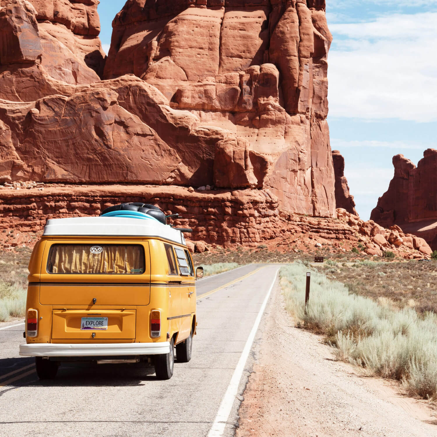 A yellow vintage van travels down an open road through a desert landscape with large red rock formations in the background. The van, embodying the spirit of summer adventure, has surfboards strapped to the roof and a blue sky with scattered clouds above. The license plate reads "EXPLORE.